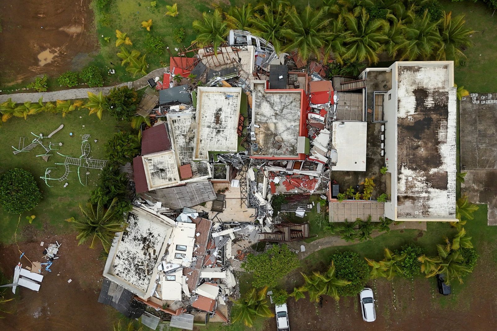 Aerial view of a cluster of buildings reduced to rubble, surrounded by palm trees.