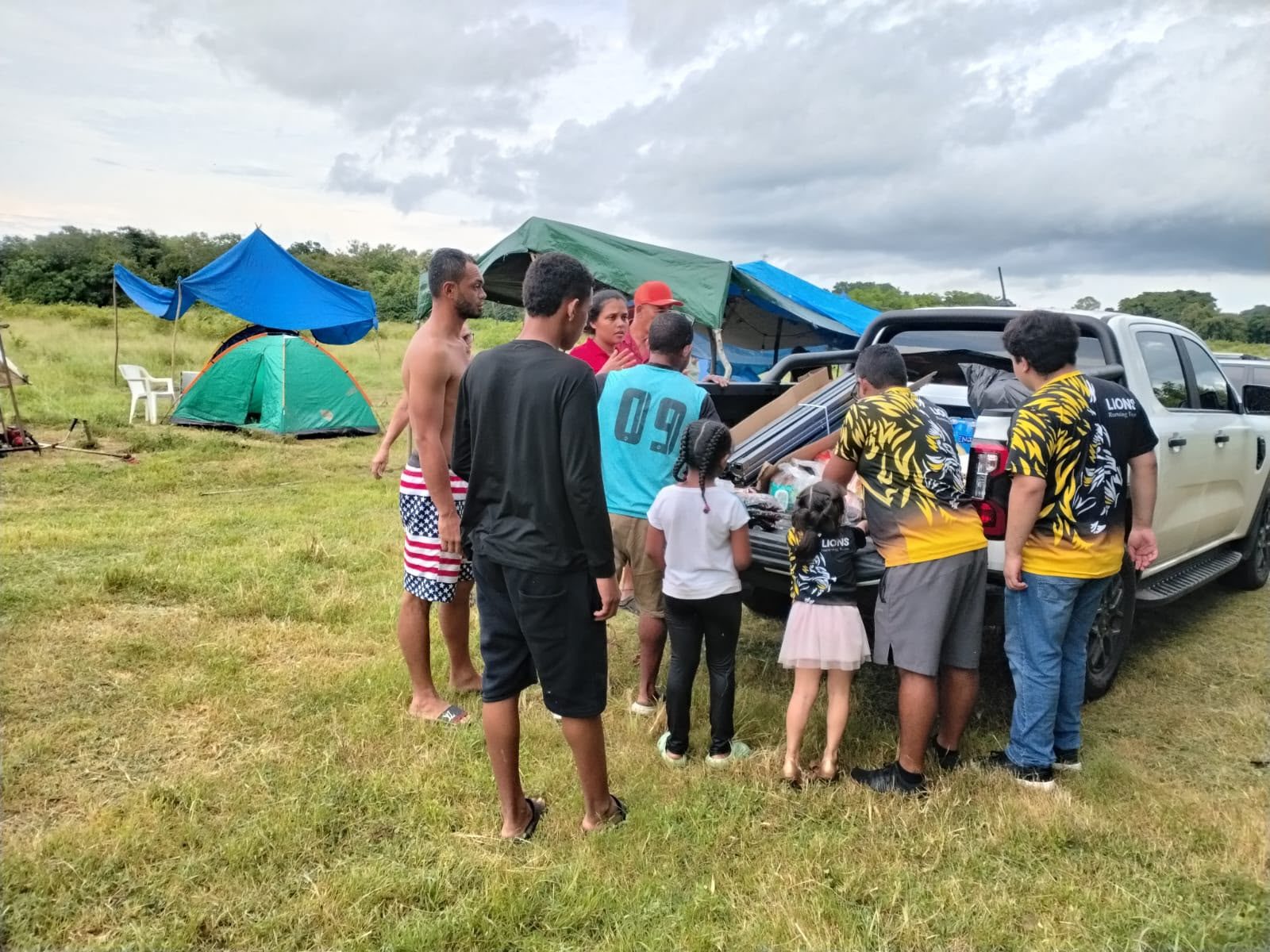 Families beside tents and tarpaulins in a field gather at a pickup truck as volunteers unload supplies.