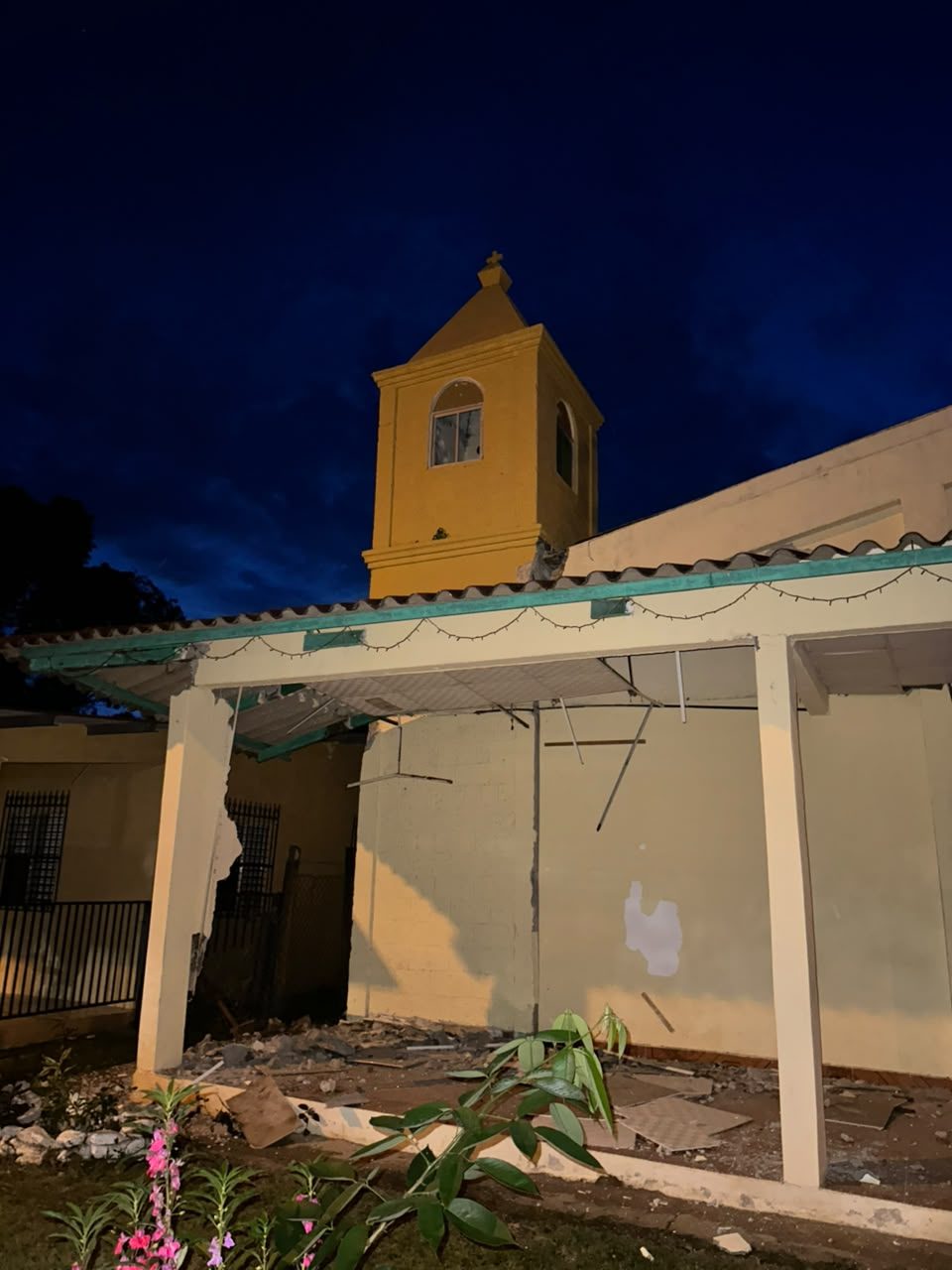 A yellow church bell tower at dusk above a porch whose wall and ceiling have been torn open, with rubble on the floor.
