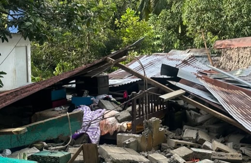A house collapsed into a pile of concrete blocks and corrugated roof sheets, with a wooden cot and bedding caught in the debris.