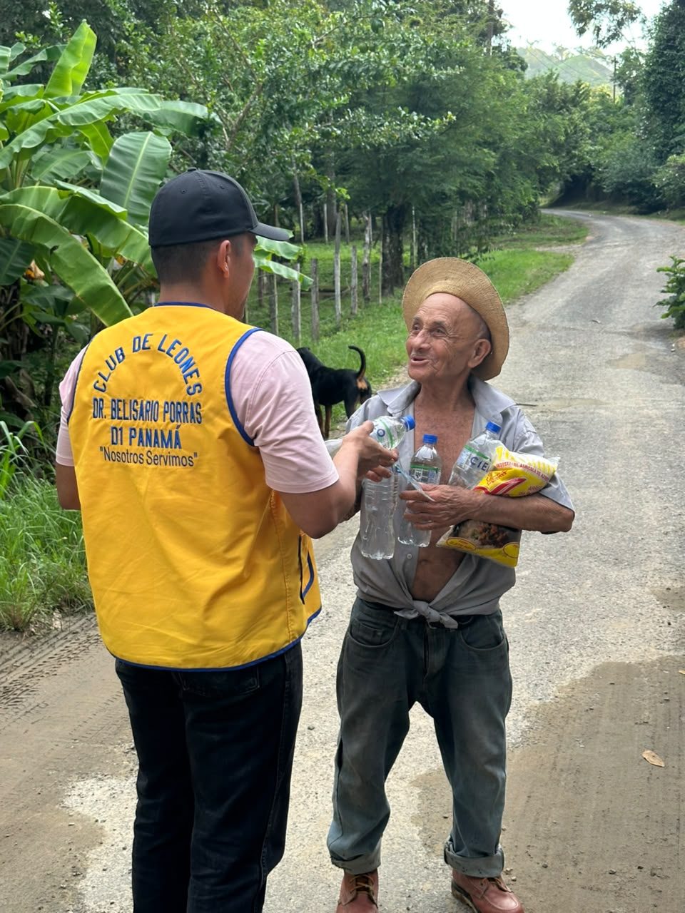 A volunteer in a Lions Club vest hands bottled water to an elderly man in a straw hat, who is already holding water and a bag of rice, on a country road.