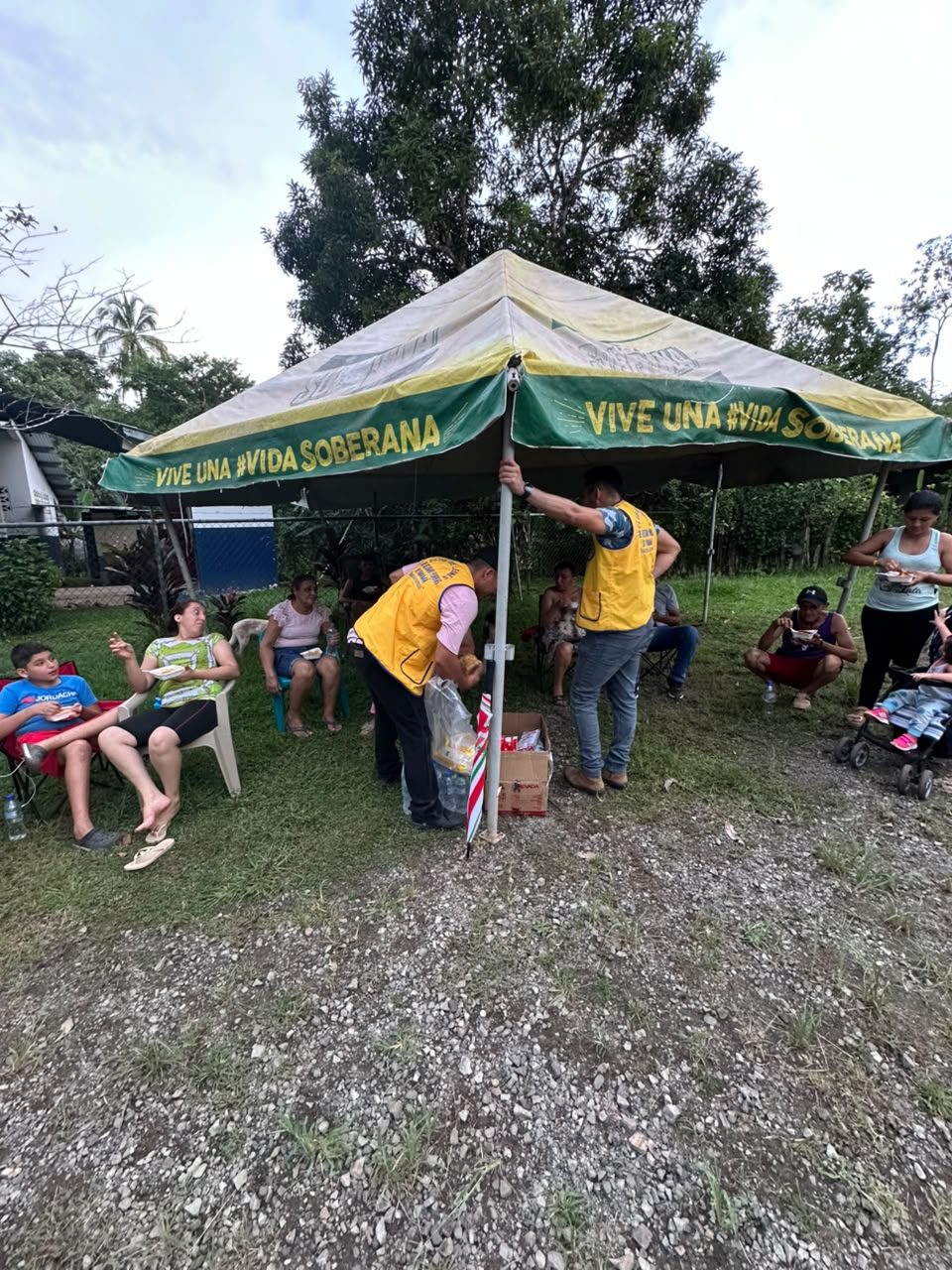 Volunteers in yellow vests hand out food under a gazebo while families sit and eat on plastic chairs.