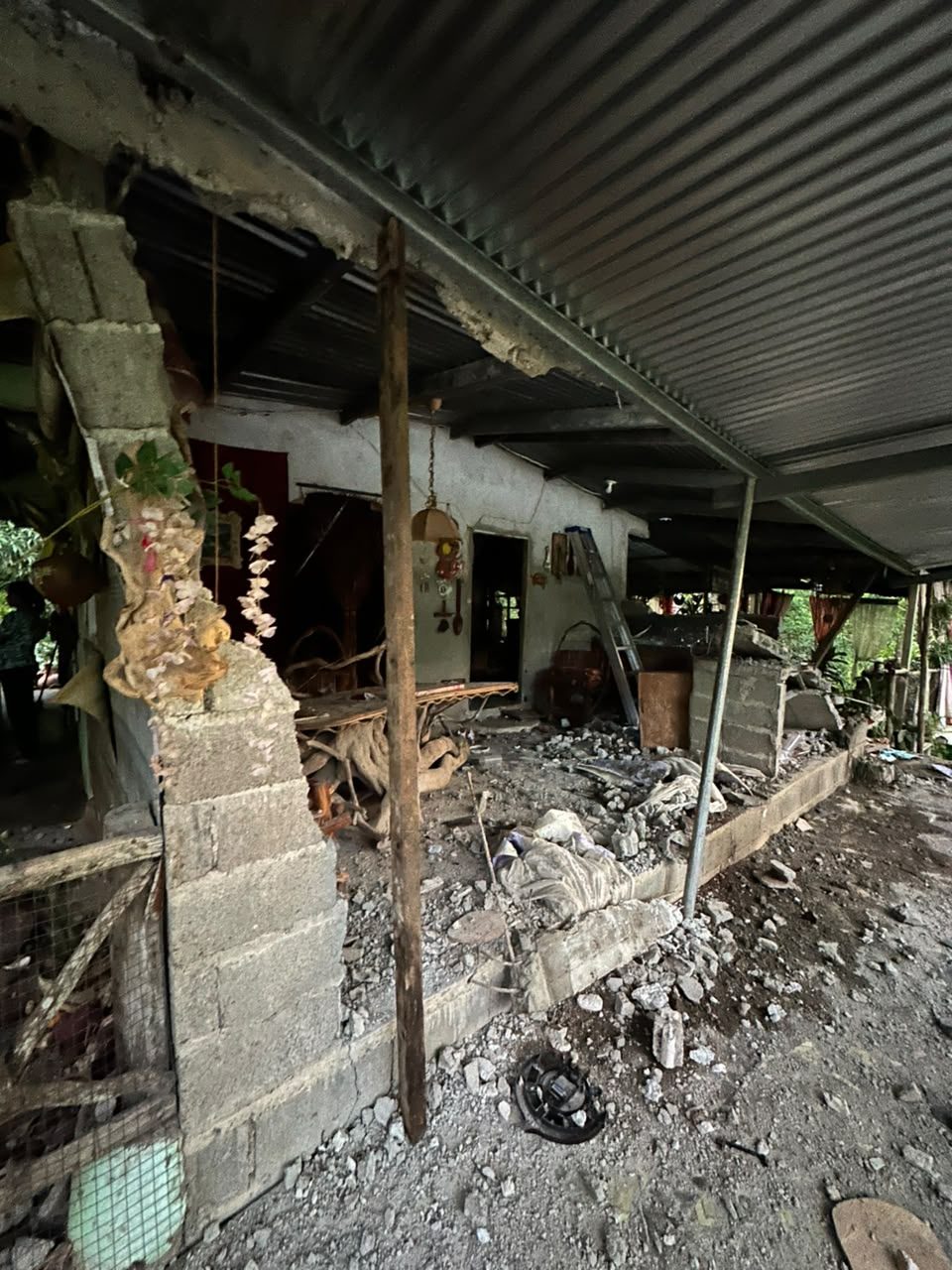 The porch of a home where concrete-block walls have broken apart and rubble covers the floor, under a metal roof propped up on a wooden pole.