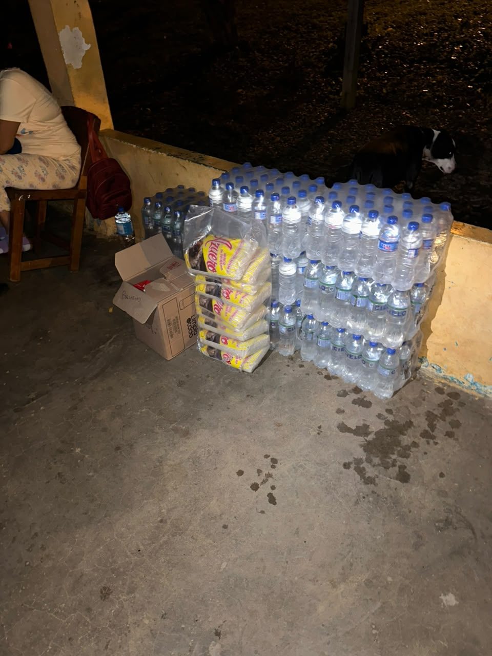 Packs of bottled water and bags of rice stacked against a low wall at night.