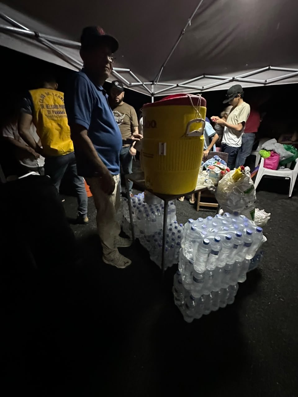 Under a gazebo at night, volunteers stand beside a water cooler and stacks of bottled water.