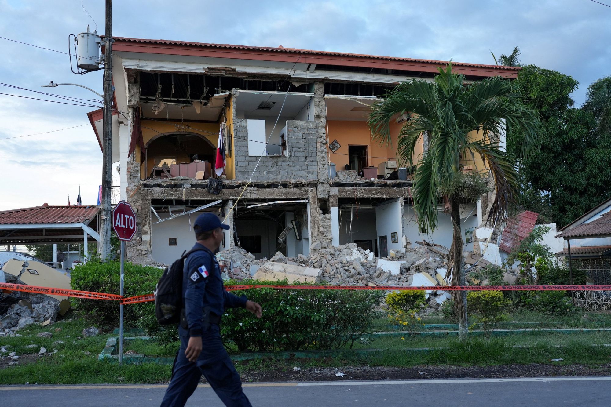 A police officer walks past a collapsed two-storey building cordoned off with civil protection tape. A Panamanian flag hangs inside a broken upstairs office.